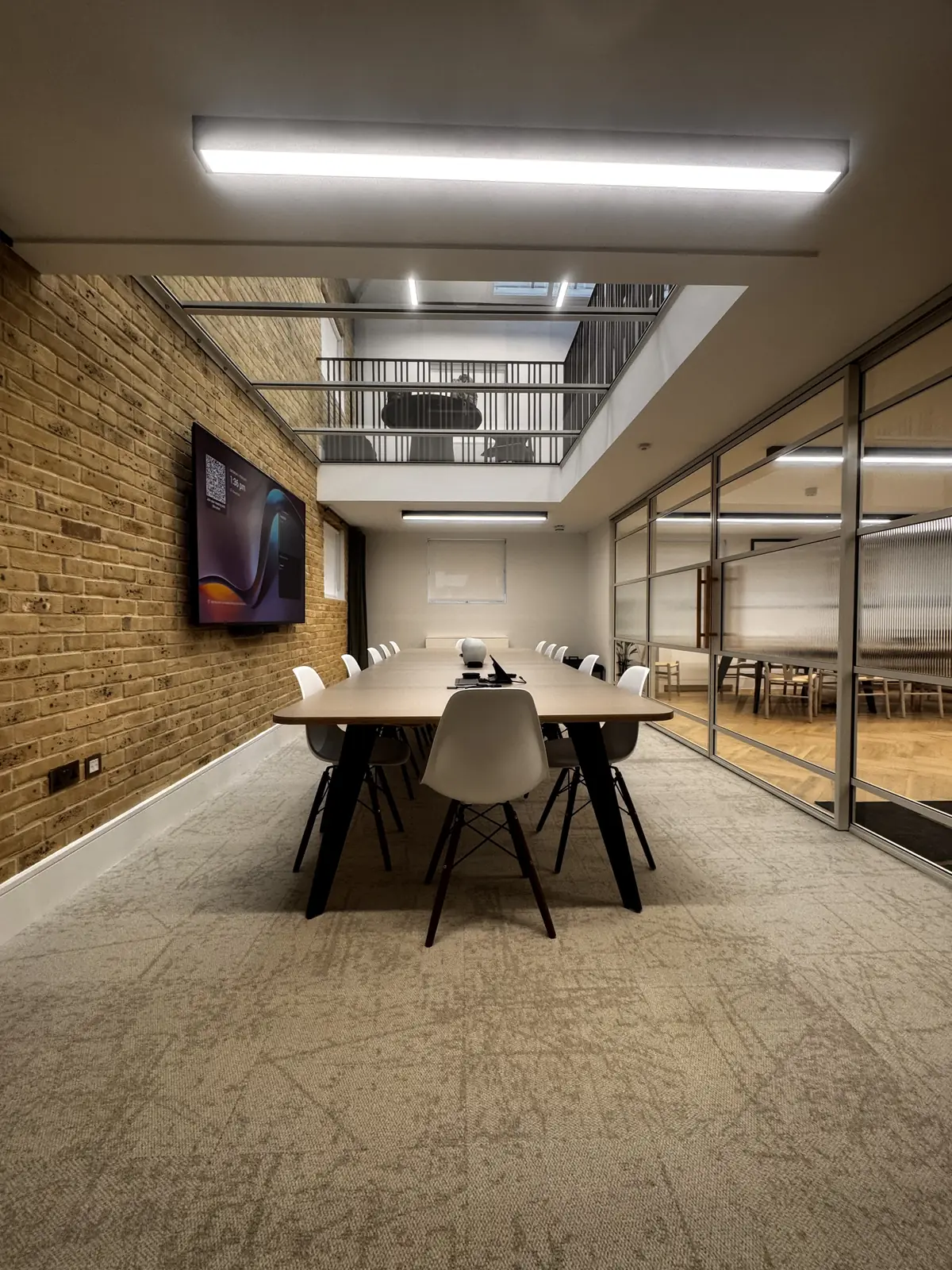KP Glazing London Modern conference room with a long table, white chairs, a wall-mounted screen, exposed brick wall, glass partitions, and an upper-level balcony visible above. Bright overhead lighting illuminates the space.