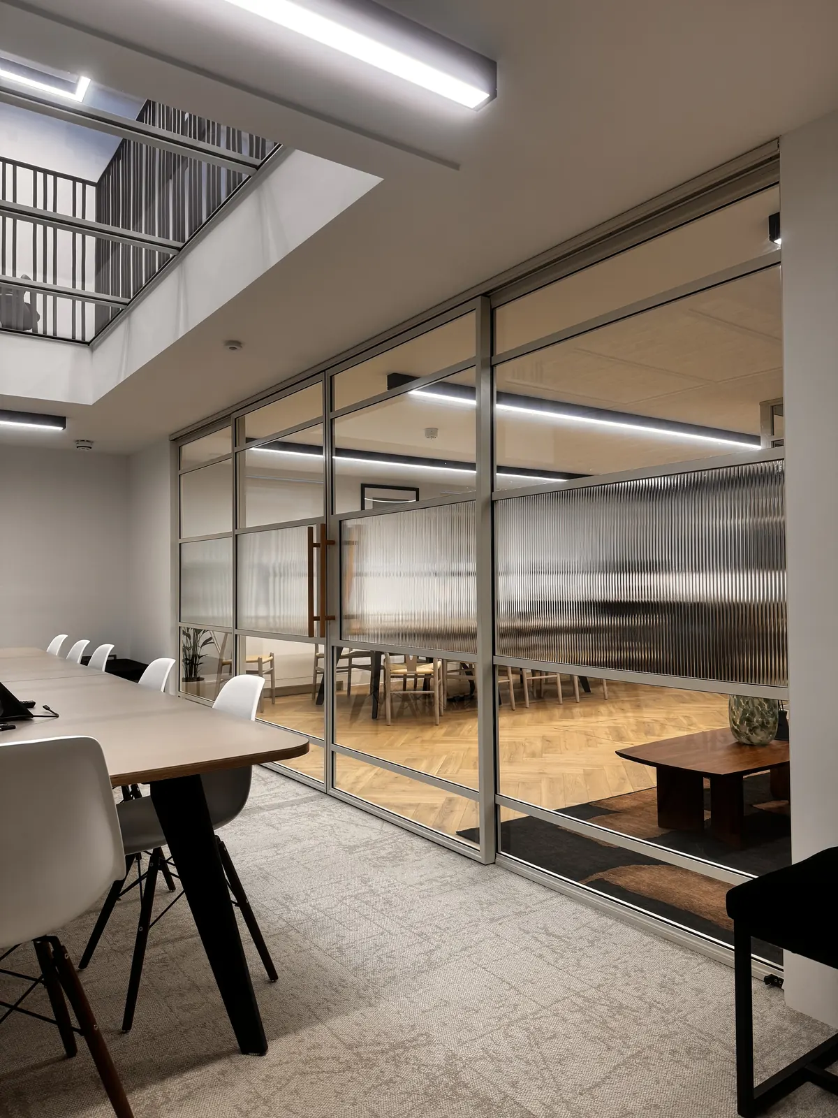 KP Glazing London Modern office space with a conference table and chairs in the foreground, glass walls separating another meeting area with wooden furniture, and natural light from an upper floor window.