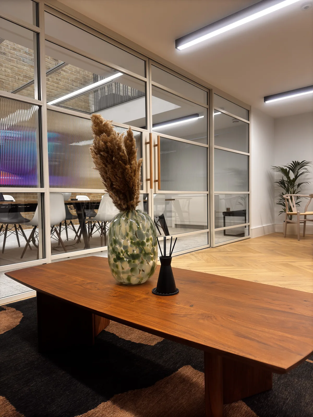 KP Glazing London A modern office lounge with a wooden coffee table, a vase of dried grasses, and a reed diffuser. Glass walls reveal a meeting room with chairs and a neon light in the background.