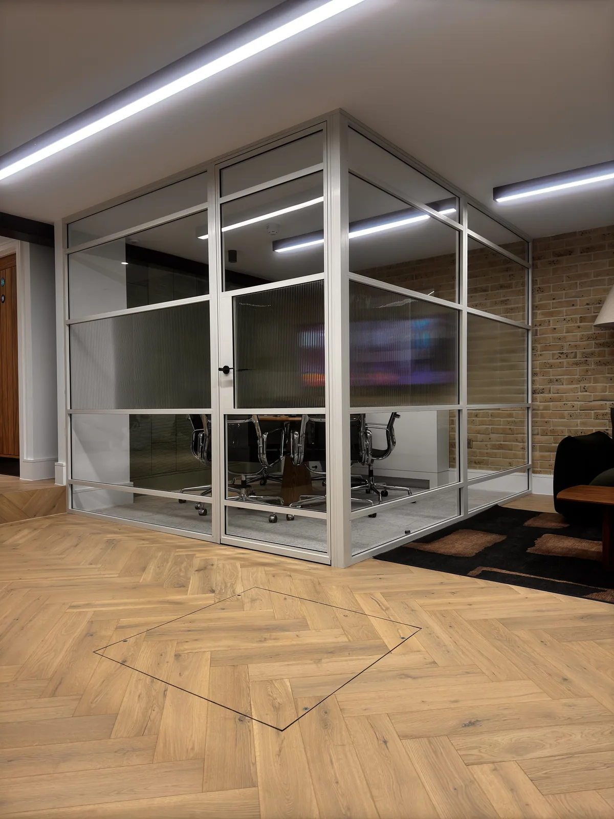KP Glazing London A modern glass-walled meeting room with black chairs around a table, located in an office with wooden flooring and exposed brick walls.
