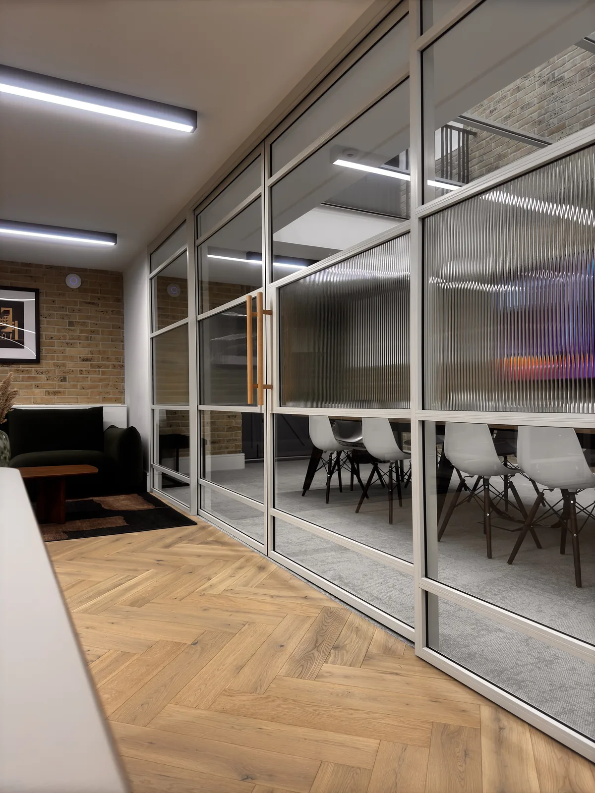 KP Glazing London Modern office space with glass walls, a long conference table with white chairs, wooden flooring, brick wall, and a seating area with a dark sofa and framed art. Fluorescent ceiling lights illuminate the area.