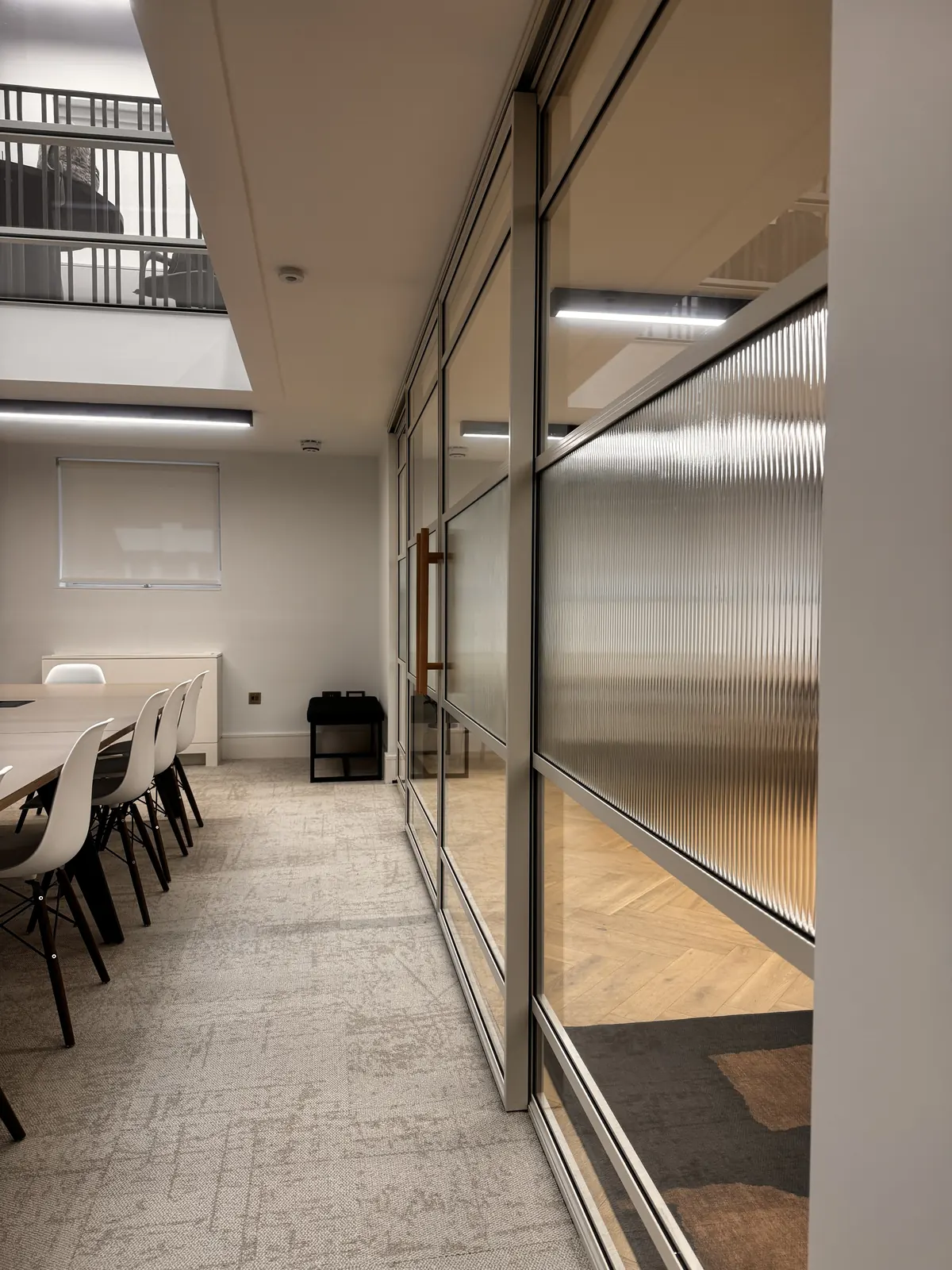 KP Glazing London Modern office space with white chairs around a long table, beige carpet, and a glass partition wall separating two rooms. Light floods in from above, highlighting the clean, minimal design.