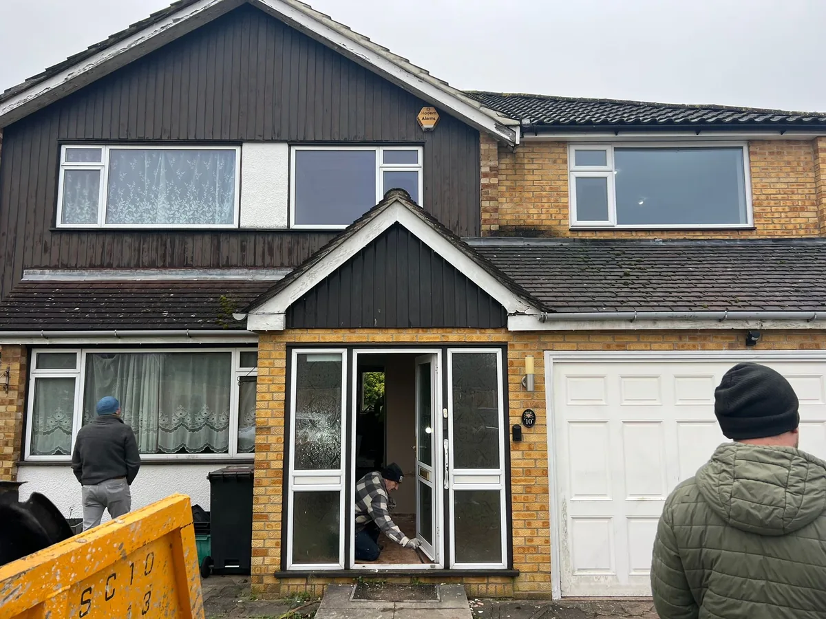 KP Glazing London A person kneels to work on the front door of a two-storey house with brown and white exterior; two other people stand nearby.
