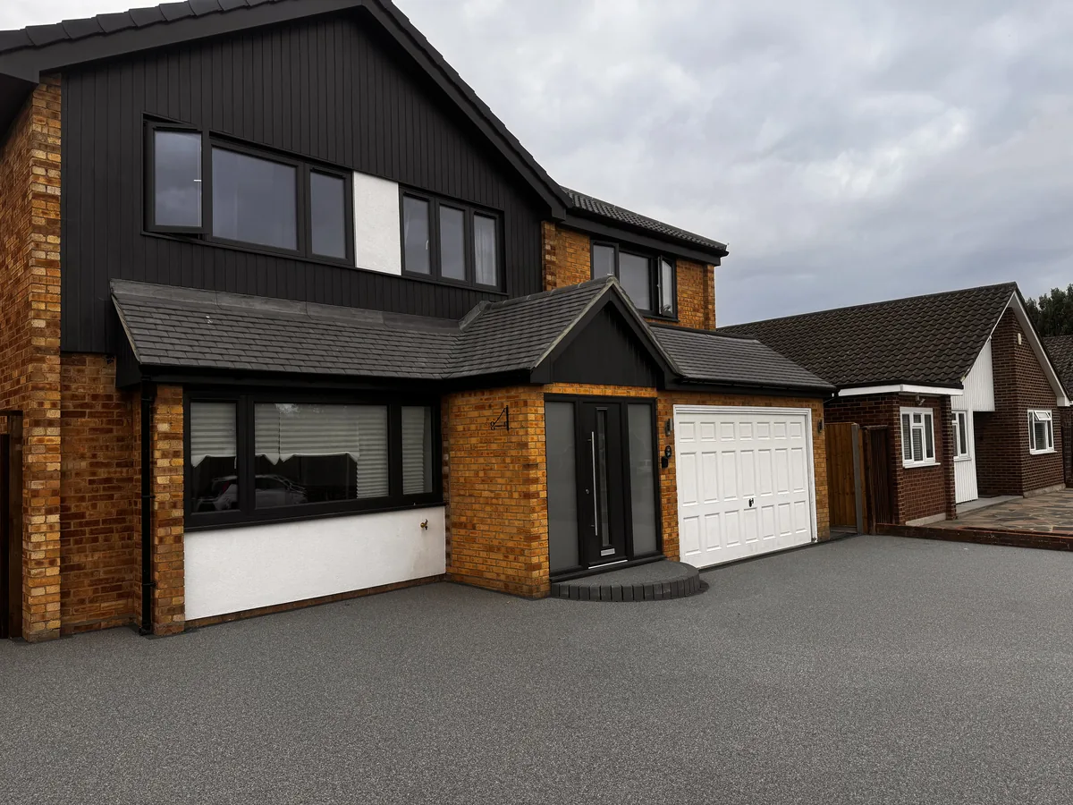 KP Glazing London Modern two-storey house with a mix of brown brick and black trim, large windows, double front doors, a white double garage, and grey driveway, alongside a single-storey brick house. Cloudy sky above.