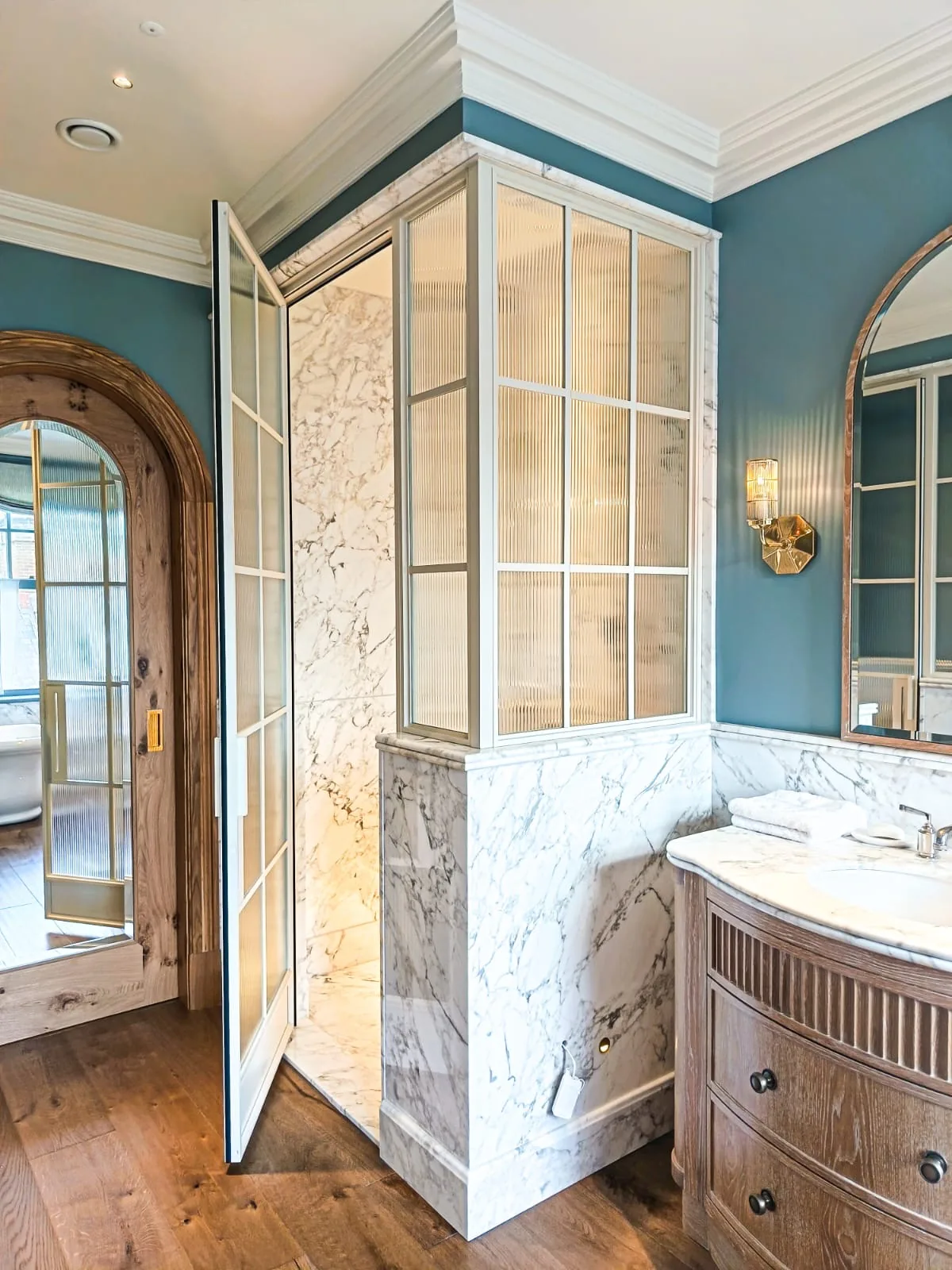 KP Glazing London Bright bathroom with blue walls, marble accents, a wooden vanity with a white basin, a glass-enclosed shower with marble walls, and wooden floors. An arched wooden door and round mirror are visible.