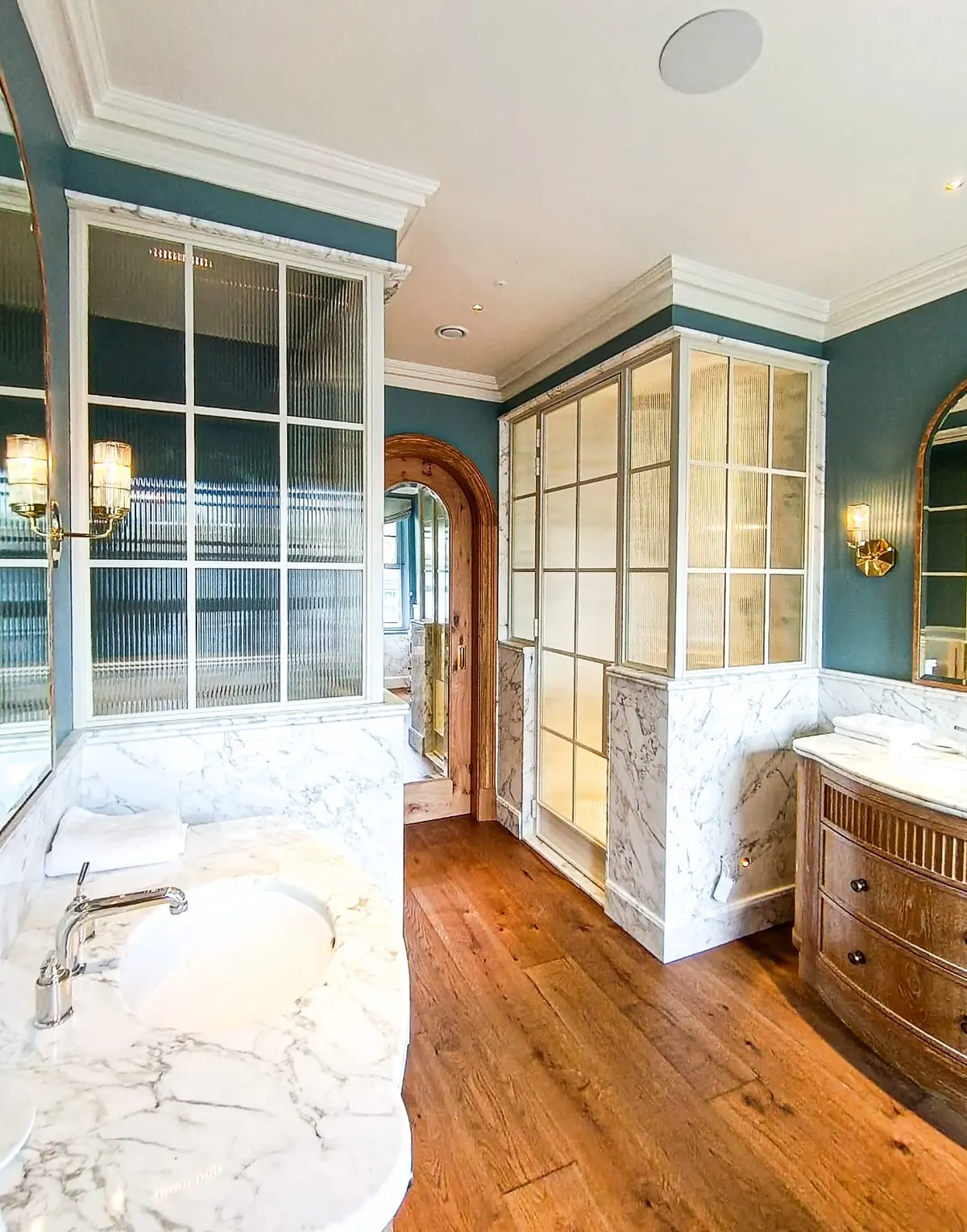 KP Glazing London Elegant bathroom with marble worktops, wooden floors, glass-panelled shower, blue walls, and gold wall lights. A wooden door and mirrors add warmth and depth to the bright, stylish space.
