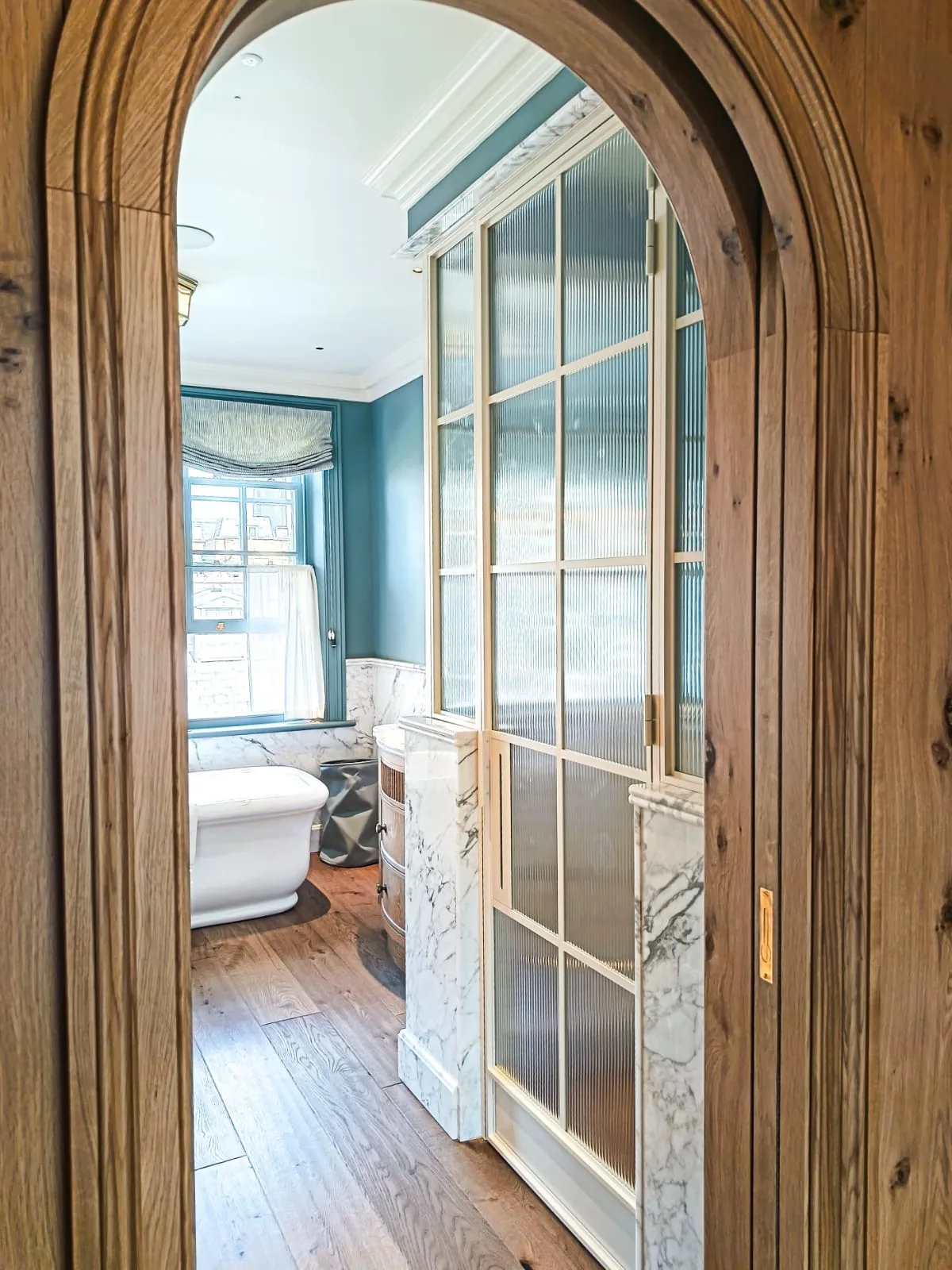 KP Glazing London A wooden arched doorway opens to a bathroom with a freestanding bath, marble accents, wooden floors, a large window with a blind, and frosted glass shower doors.
