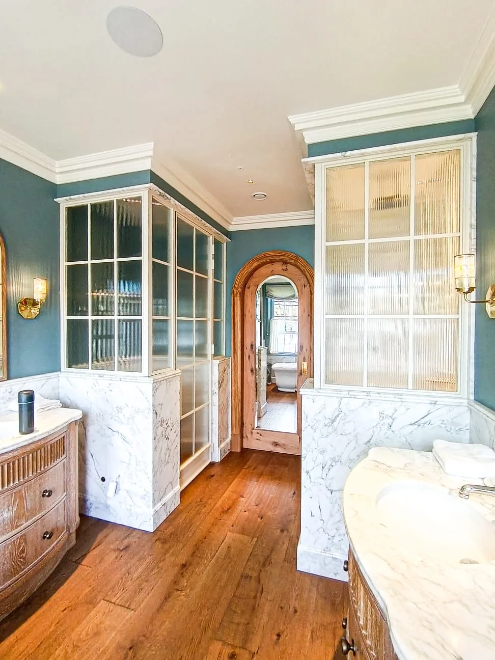 KP Glazing London A bathroom with blue-green walls, marble surfaces, wooden flooring, glass-panelled dividers, a wooden arched door, and brass light fittings. The vanity has a curved marble worktop and a towel on it.