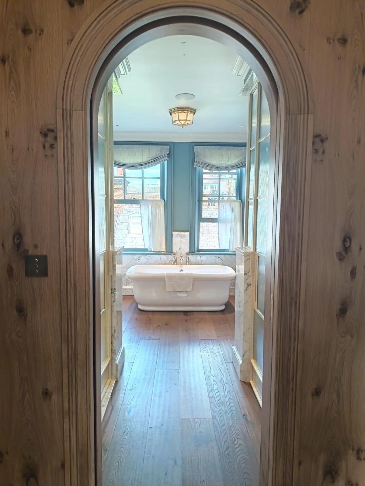KP Glazing London View through a wooden arched doorway into a bathroom with a white freestanding bath, large windows with sheer curtains, and wooden floors. Natural light fills the room.