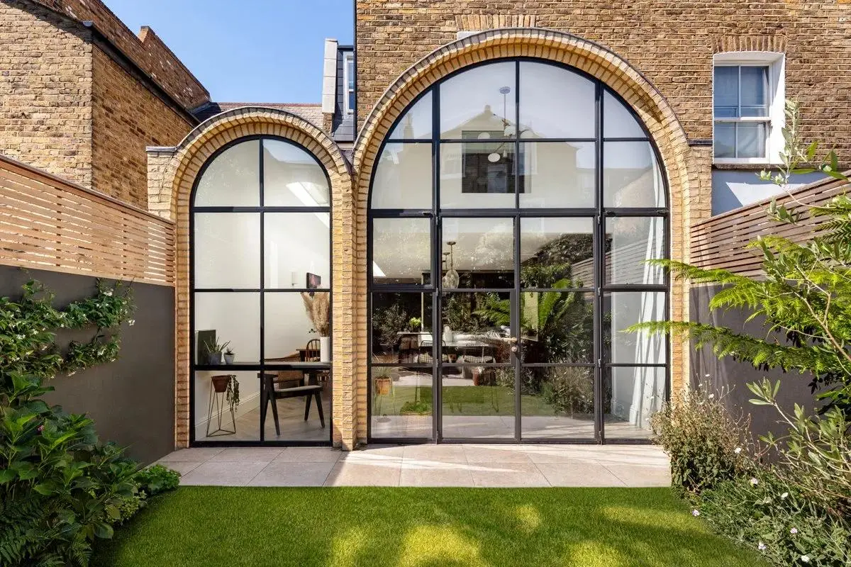 KP Glazing London Exterior view of a modern house with large arched windows, brick walls, and a lush green lawn. Reflections in the glass show the garden, and there is patio space with outdoor plants on either side.
