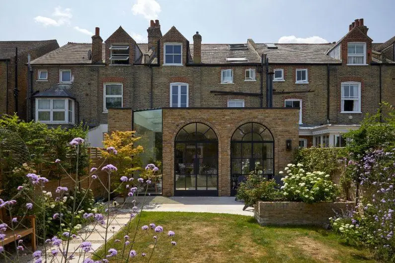 KP Glazing London Back garden view of a brick terraced house with large arched windows and doors, surrounded by flower beds with flowers and greenery under a blue sky.