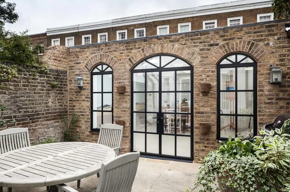 KP Glazing London A brick patio features a round wooden table with chairs, potted plants, and arched glass doors leading into a bright, modern interior.