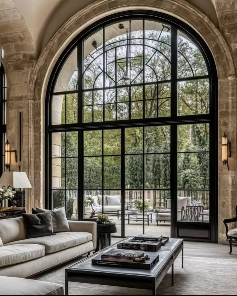 KP Glazing London Elegant living room with arched, black-framed windows, beige sofas, and a coffee table. Outdoor patio with seating is visible through the large glass doors, surrounded by trees and greenery.