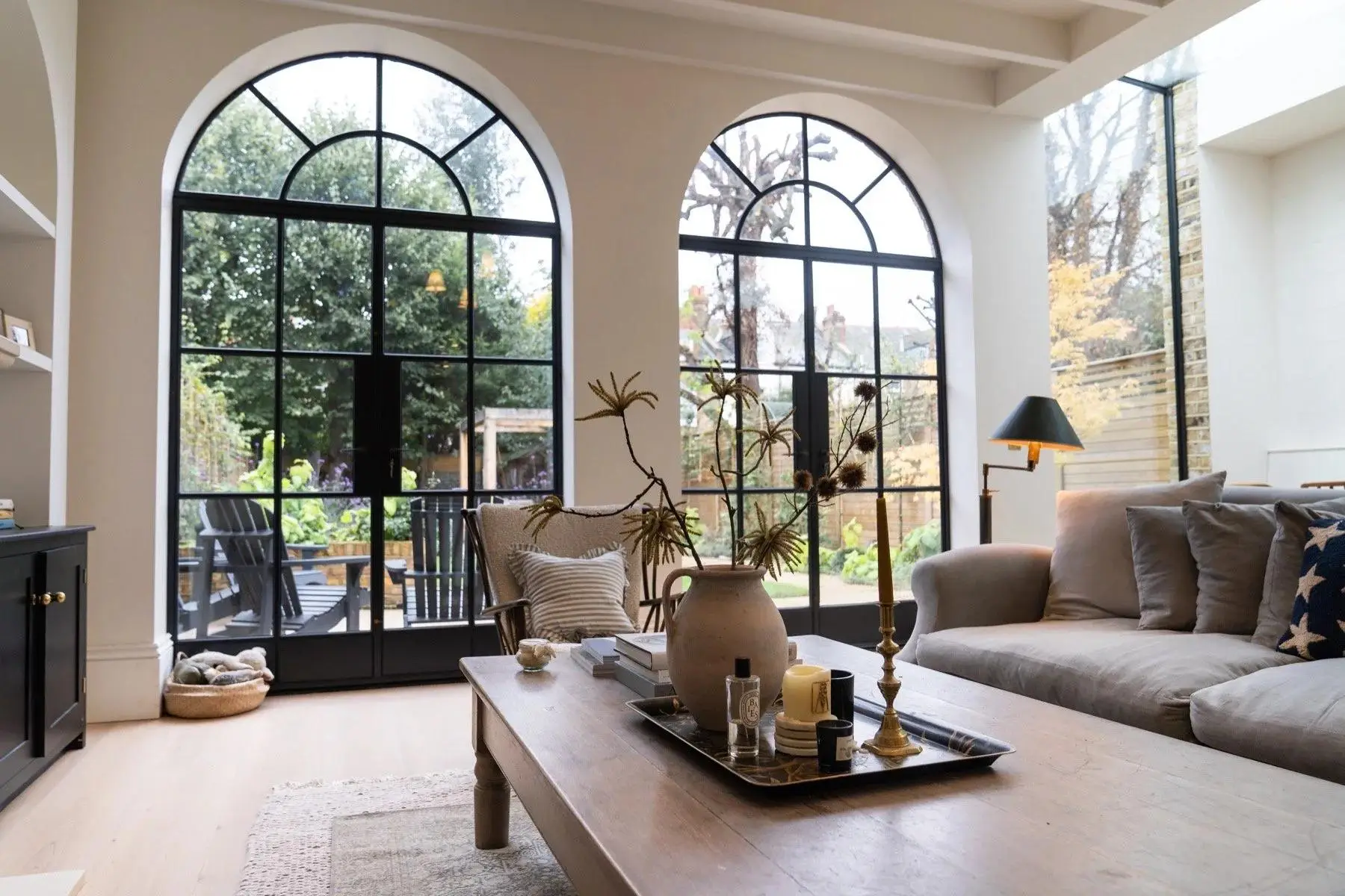 KP Glazing London A cosy living room with a sofa, coffee table, and large arched windows overlooking a garden with patio chairs. Sunlight streams in, creating a bright, inviting atmosphere.