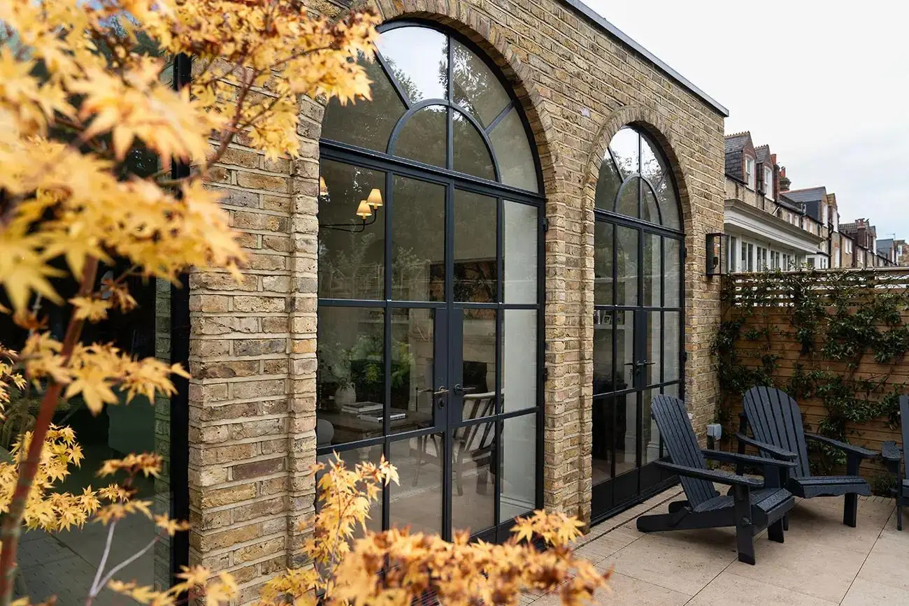 KP Glazing London Brick house with large arched glass doors, two black Adirondack chairs on a patio, and a tree with yellow leaves in the foreground.