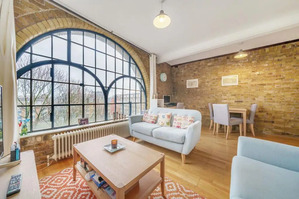 KP Glazing London A cosy living room with exposed brick walls, arched industrial window, light wood floors, pastel sofas, a wooden coffee table, dining area, wall decor, pendant lights, and a patterned orange rug.