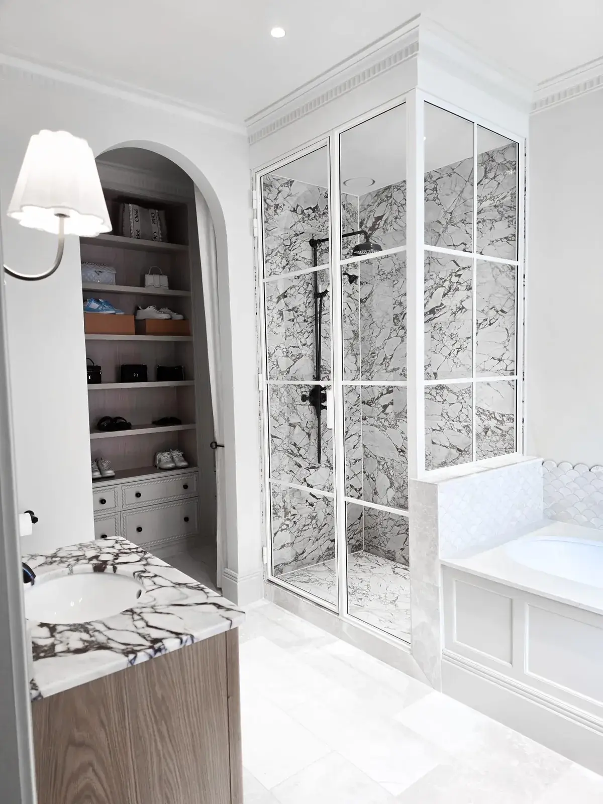 KP Glazing London A bathroom featuring a marble-topped vanity unit, a glass-enclosed shower with marble walls, and an arched doorway leading to a walk-in wardrobe with built-in shelving.