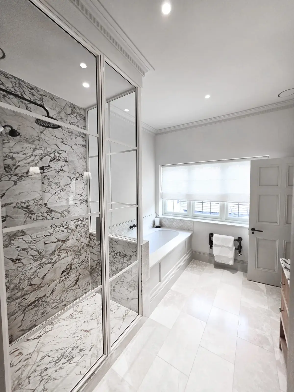 KP Glazing London A bright bathroom featuring a walk-in shower with white-and-grey marble tiling, a built-in bath under a window, and a white door.