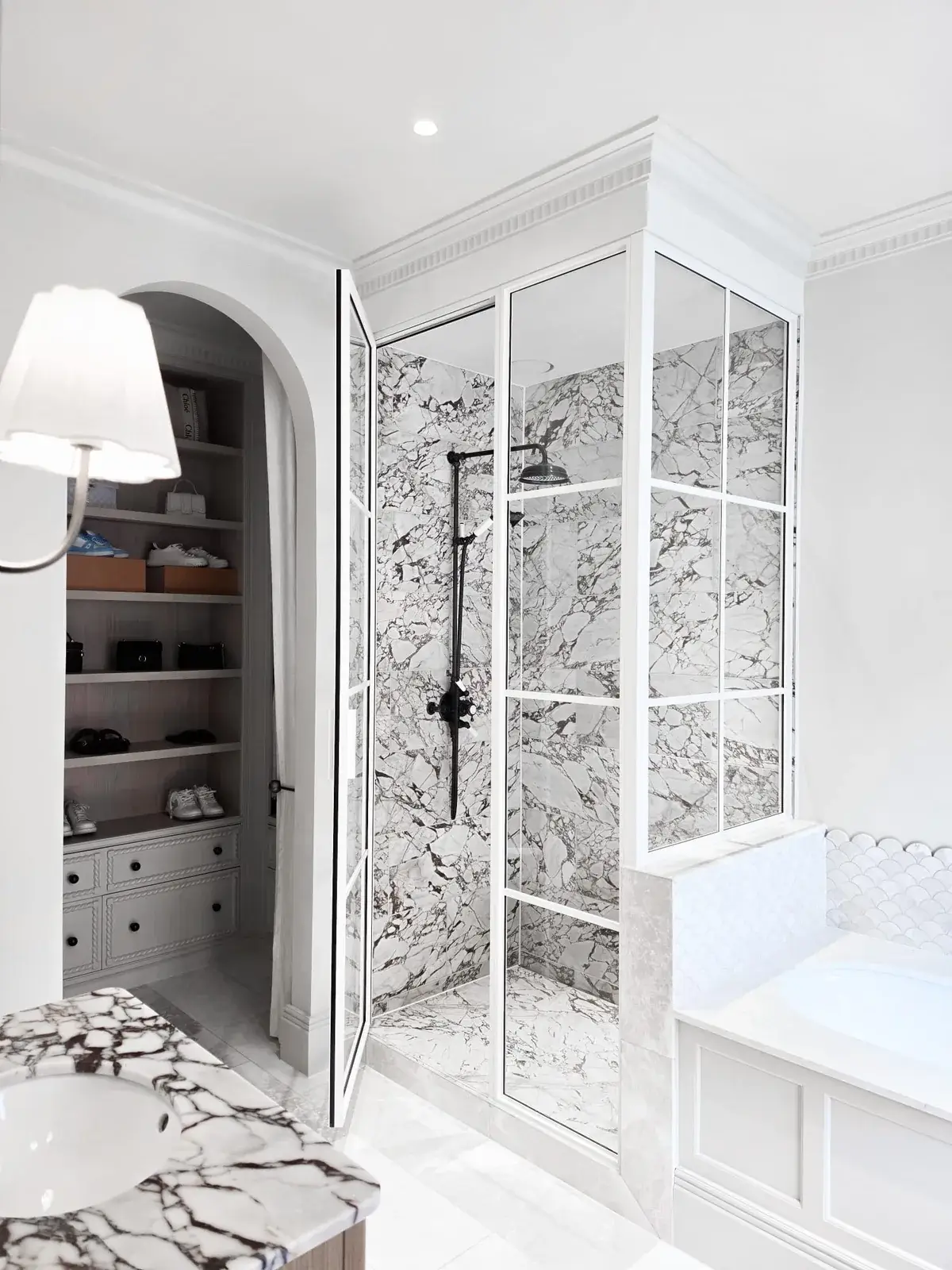 KP Glazing London A glass-enclosed shower with black metal framing and marble walls, situated next to an arched walk-in wardrobe and a white bath.