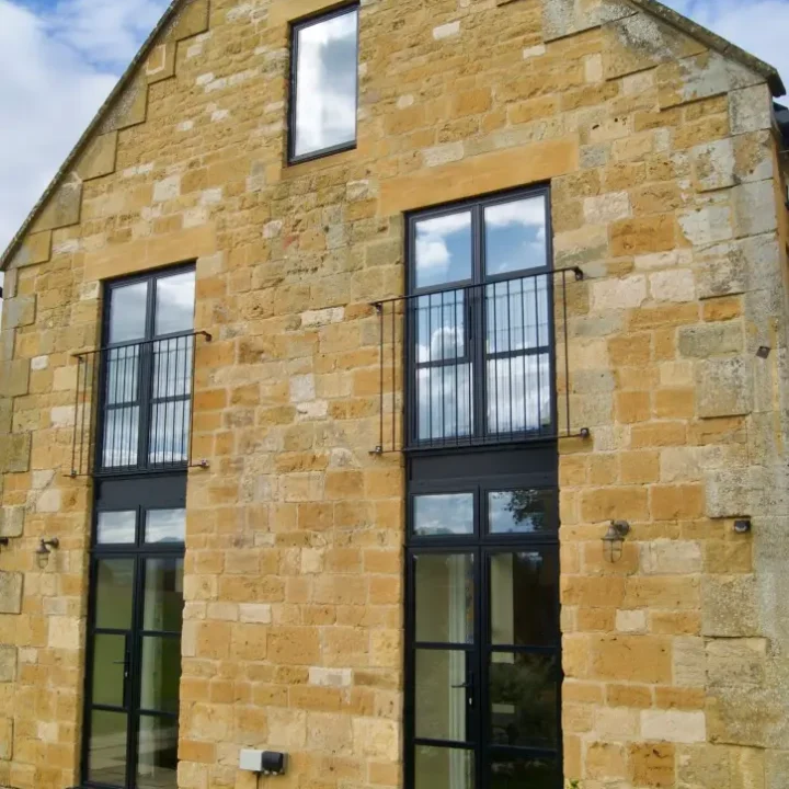 KP Glazing London A modernised stone barn conversion with large black-framed windows and a high gable roof, set against a blue sky with white clouds. There are flowers and paving stones in the foreground.