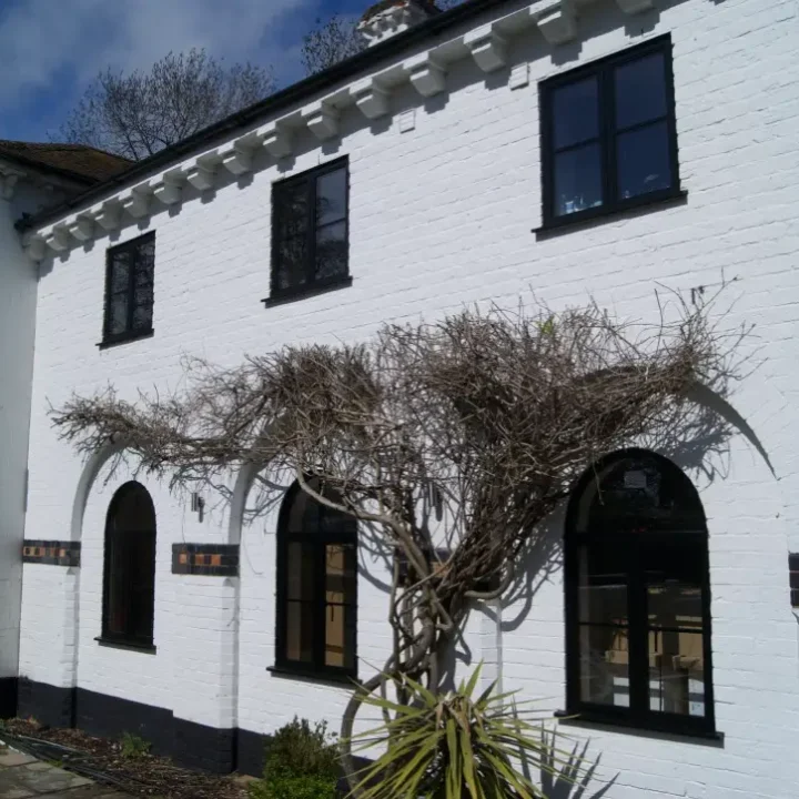KP Glazing London A white brick house with black-trimmed windows and doors, featuring bare, twisting branches growing across the front façade, set against a blue sky with some clouds.
