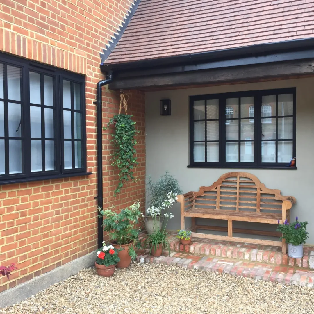 KP Glazing London A wooden bench sits on a brick porch outside a house with red brick walls. Potted plants and hanging greenery decorate the entrance, and gravel covers the ground in front.