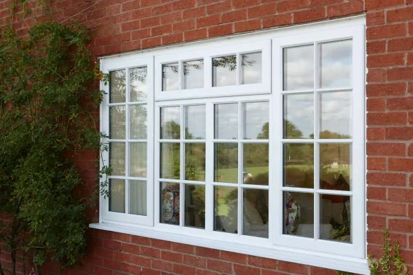 KP Glazing London A white-framed window set in a red brick wall, reflecting trees and a blue sky outside. Green foliage climbs up one side of the wall next to the window.