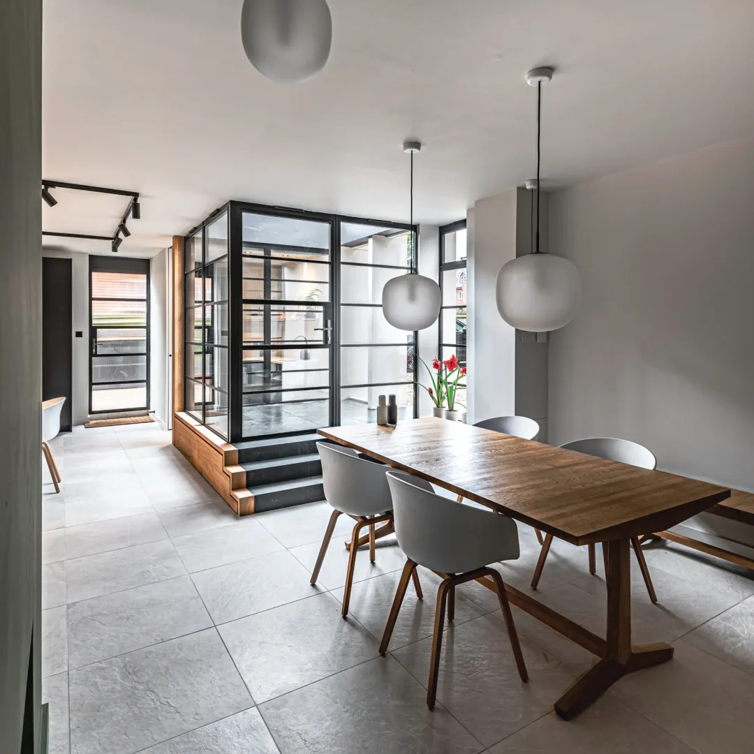 KP Glazing London Modern dining room with a wooden table, white chairs, large pendant lights, tiled floor, and glass walls; a vase with red flowers sits on the table near windows letting in natural light.