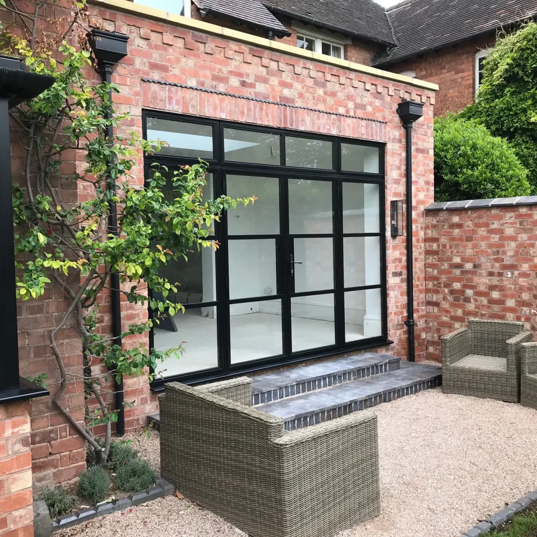 KP Glazing London A modern glass door with black frames opens from a brick building onto a small patio with wicker chairs and a sofa, surrounded by brick walls and plants.