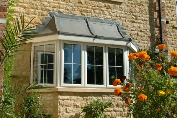 KP Glazing London A stone house features a bay window with white frames and a grey awning. Yellow-orange flowers and green plants grow nearby, with sunlight casting shadows on the wall.
