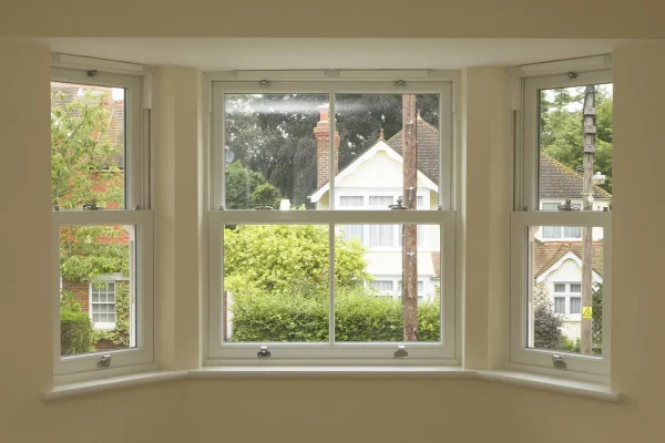 KP Glazing London View through a bay window showing three white-framed windows overlooking suburban houses, greenery, and trees outside on a cloudy day.