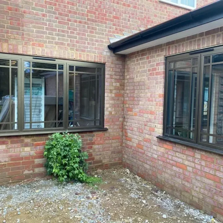 KP Glazing London A brick exterior corner of a house with two large, dark-framed windows. A small green bush grows at the base between the windows, and the ground is covered with earth and scattered gravel.